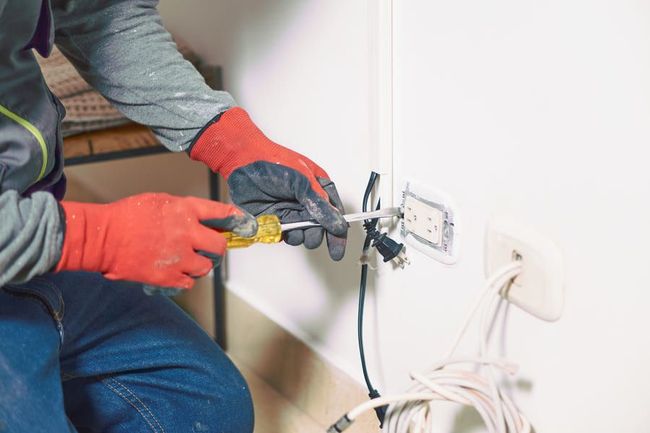Une personne portant des gants rouges utilise un outil électrique pour installer une prise électrique dans un mur.