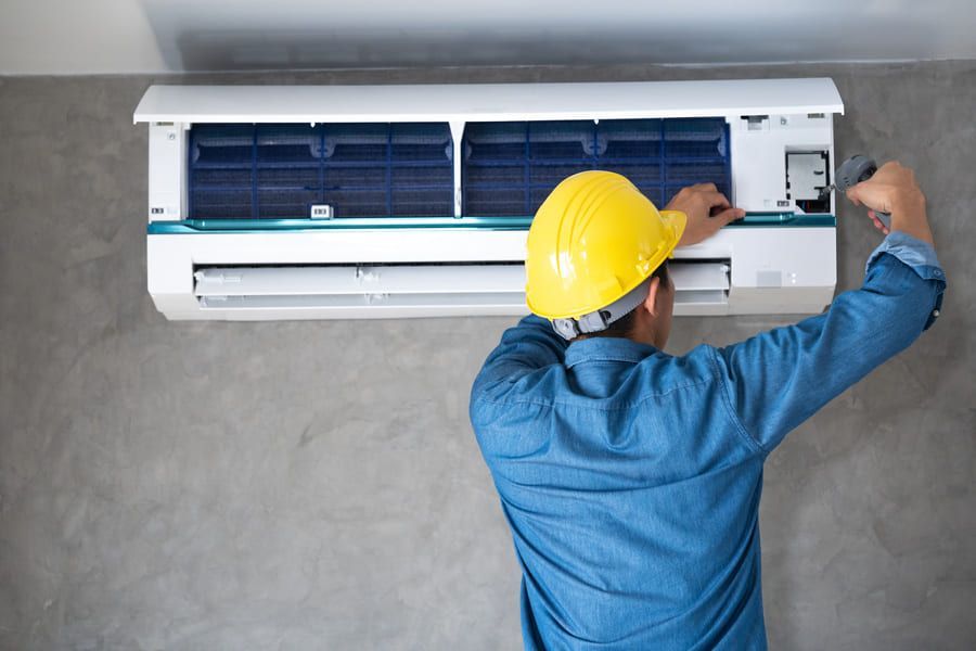 Un technicien portant un casque jaune répare un climatiseur mural à l'intérieur d'une pièce.