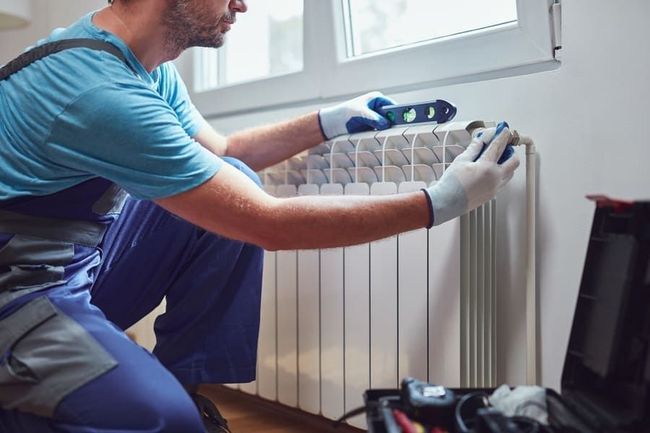 Un technicien installe un radiateur blanc sous une fenêtre à l'aide d'un niveau à bulle et d'une clé.
