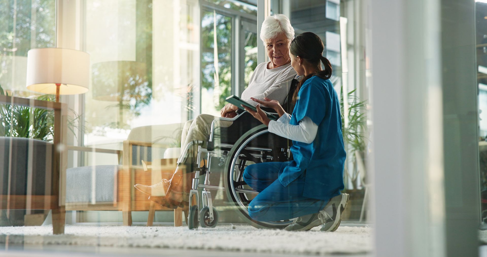 Infirmière montrant une tablette à une femme âgée en fauteuil roulant. Intérieur ensoleillé.