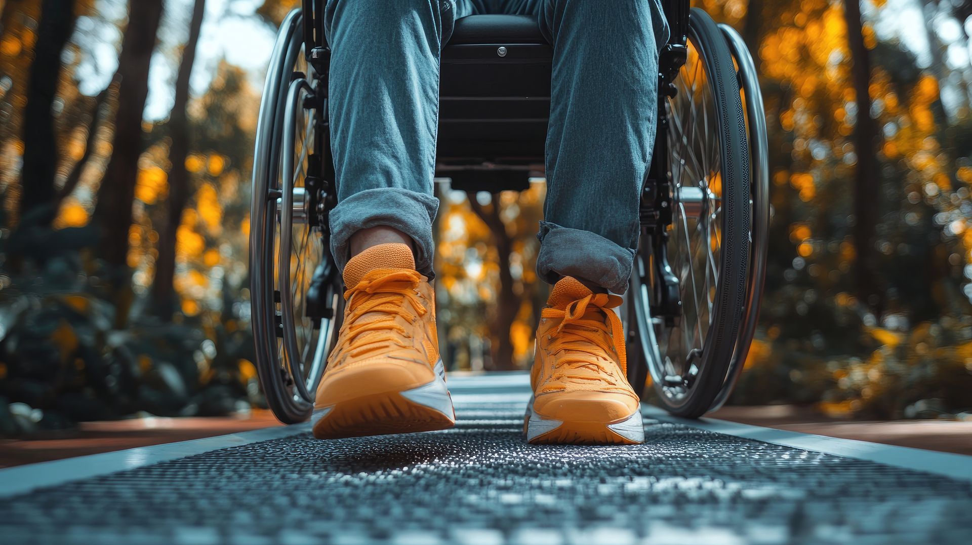 Personne en fauteuil roulant, chaussée de baskets orange, roulant sur un chemin texturé. Décor automnal avec arbres.