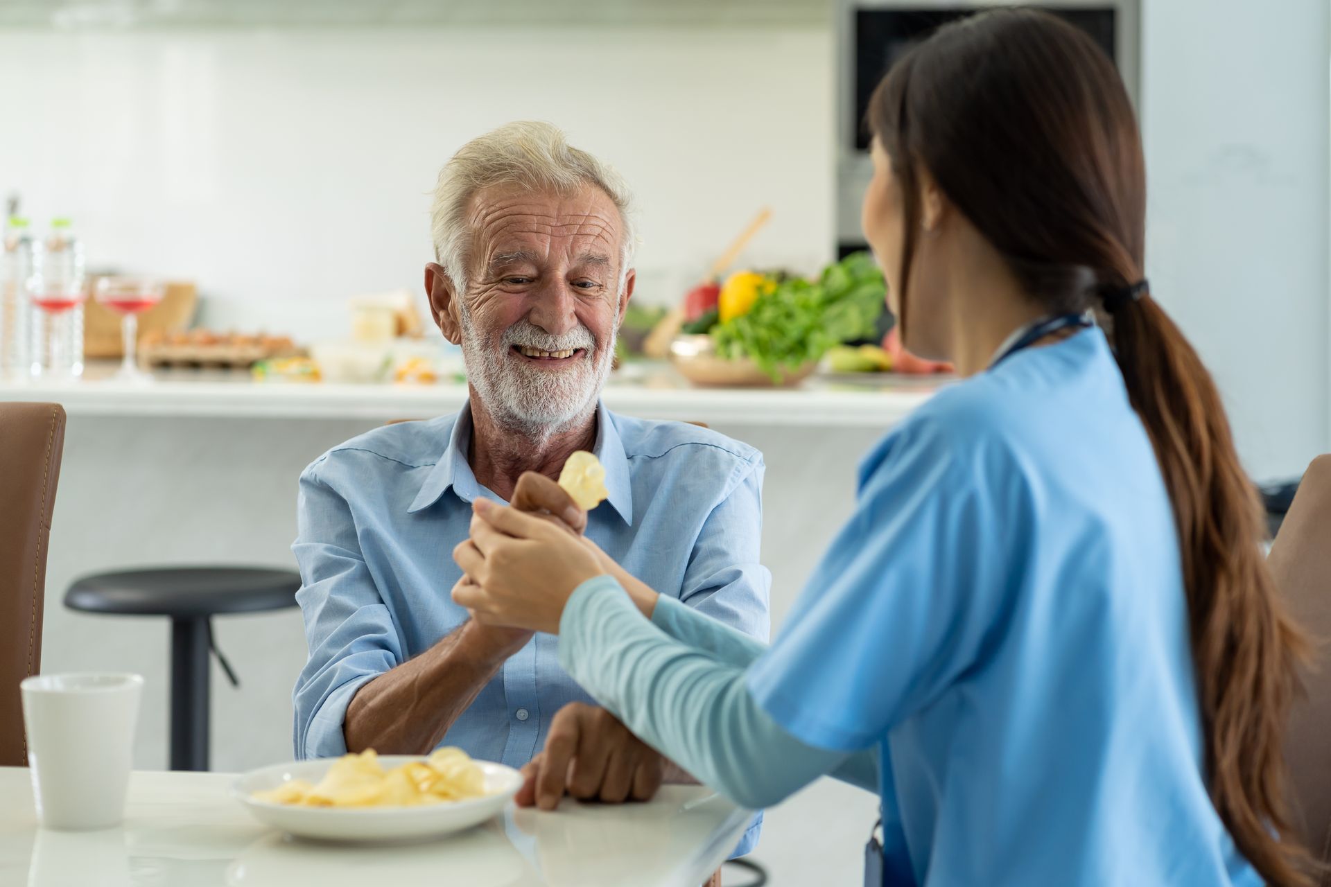Un soignant en tenue bleue offre de la nourriture à un homme âgé, souriant, assis à une table de cuisine.