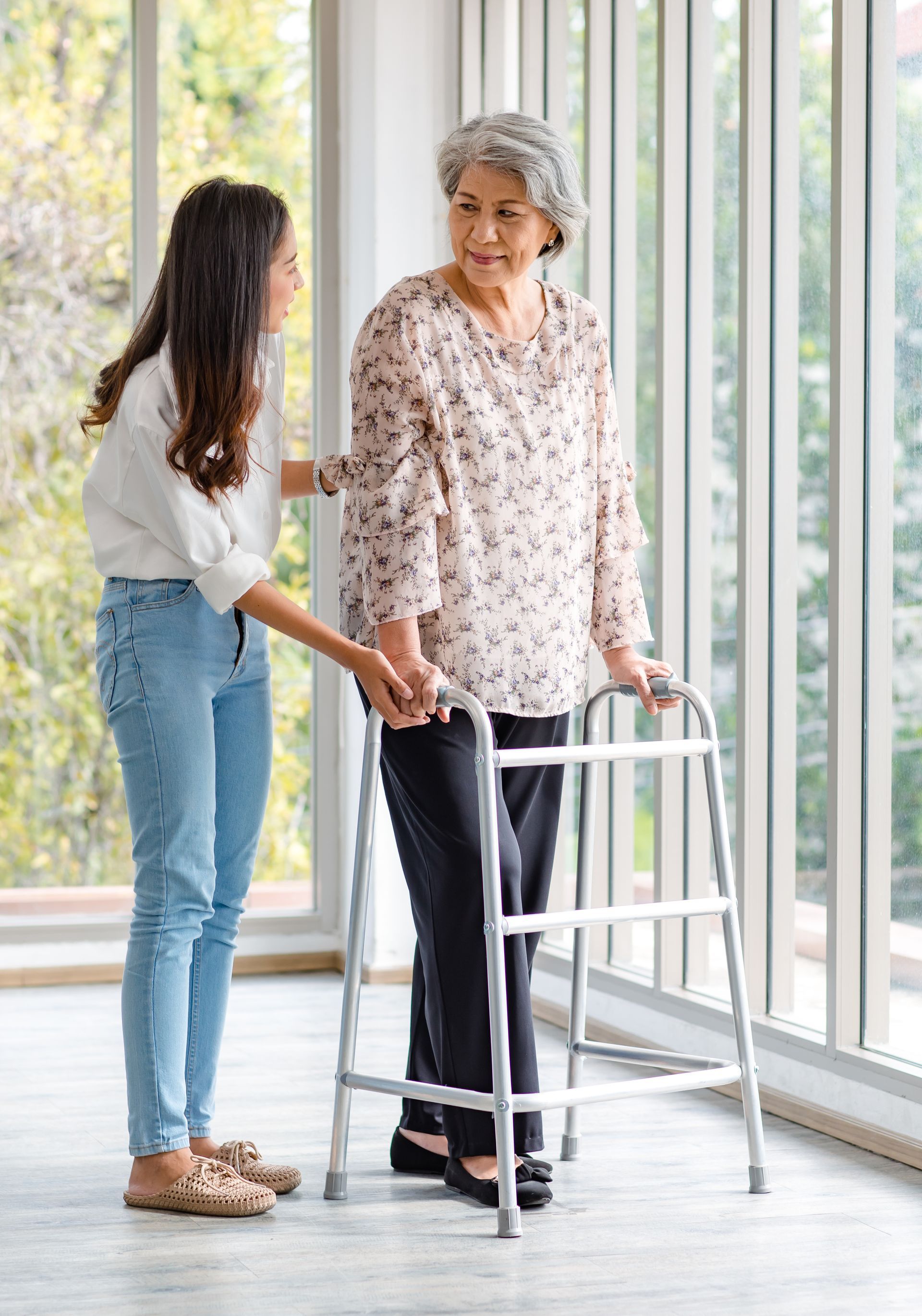 Une jeune femme aide une femme âgée à marcher avec un déambulateur près d'une fenêtre.