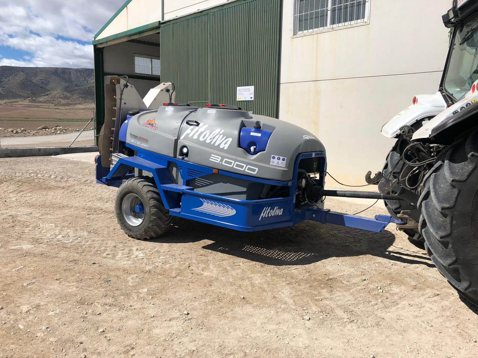 Un pulverizador azul y gris está estacionado junto a un tractor frente a un edificio.