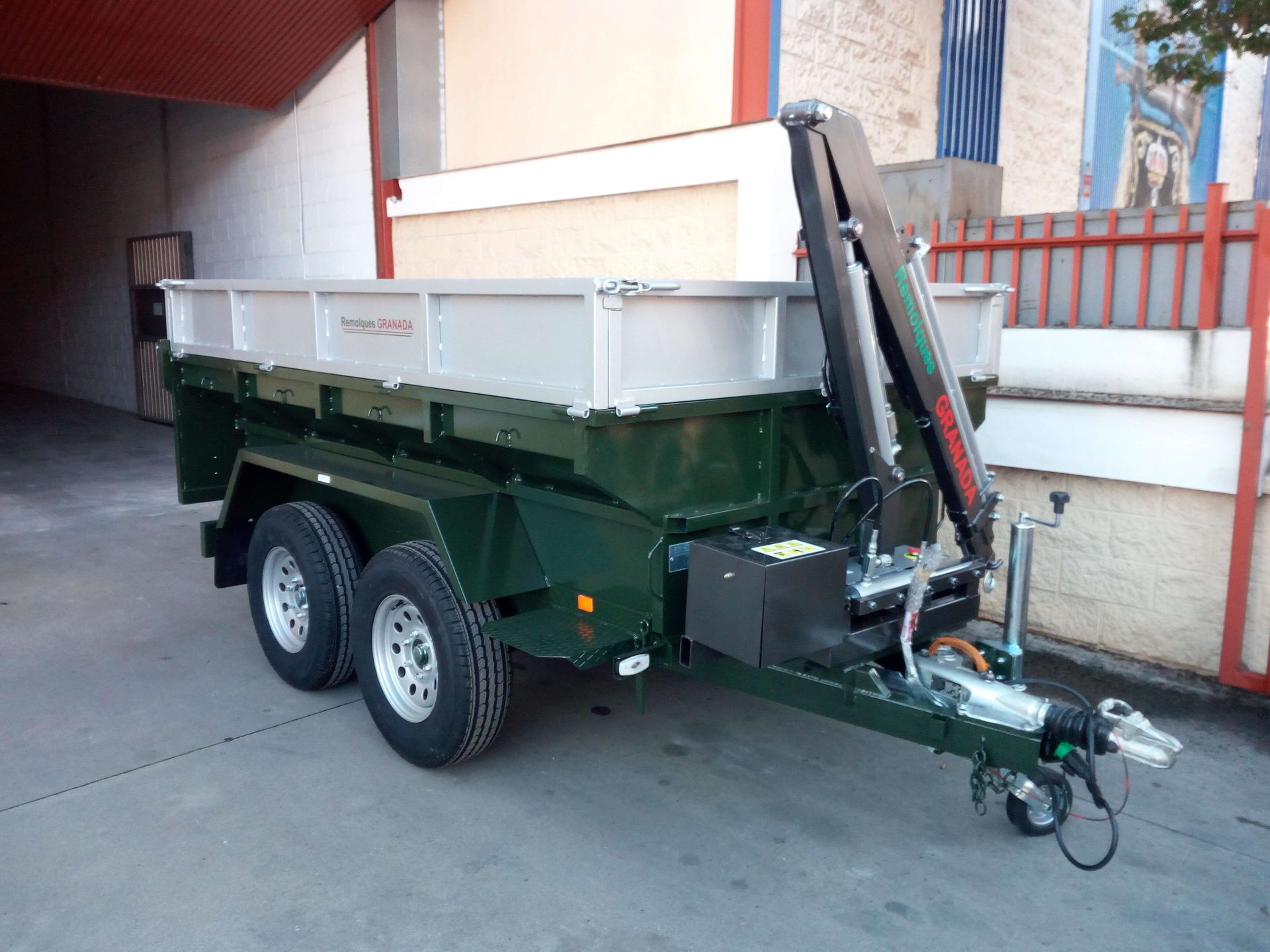 Un remolque volquete con una grúa acoplada está estacionado frente a un edificio.