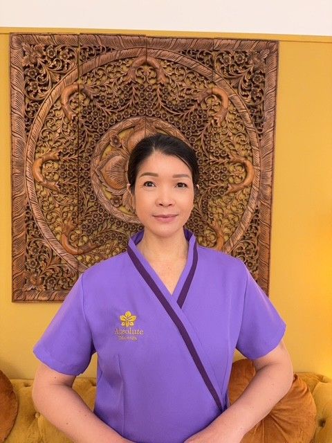 Lächelnde Frau in einem lila Oberteil, die Hände zum Gruß gefaltet. Vor einer geschnitzten Holzplatte und einer gelben Wand.