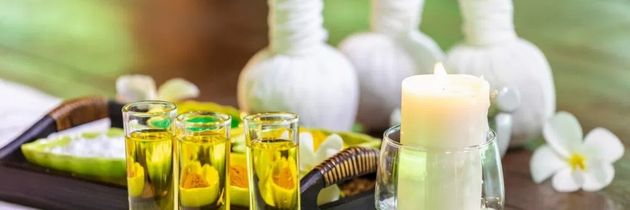 Spa-Behandlungssetup: ätherische Öle, Kerzen, Massagebälle und Blumen auf einem Tablett. Sanfte Grün- und Weißtöne.