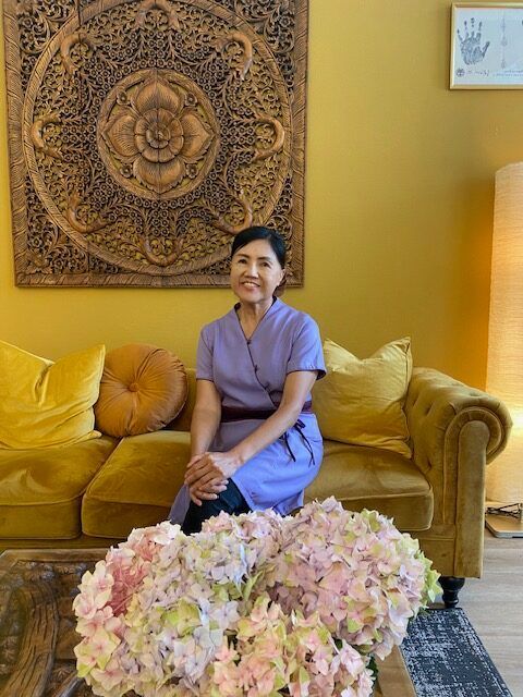Frau in blauer Uniform sitzt auf goldenem Sofa, davor Blumen, mit Holzwandkunst.