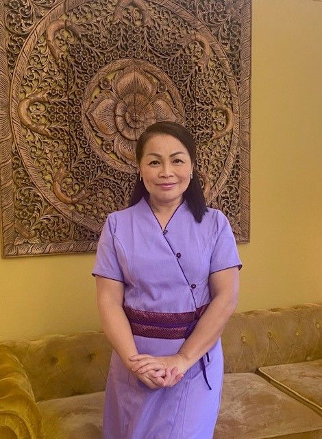 Frau in lila Thai-Massageuniform steht vor dekorativem Wandpaneel.