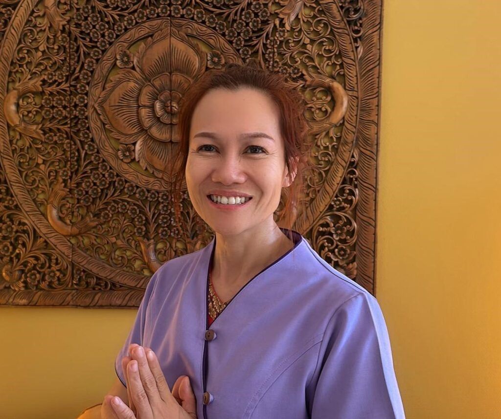 Lächelnde Frau in einem lila Oberteil, die Hände zum Gruß gefaltet. Vor einer geschnitzten Holzplatte und einer gelben Wand.