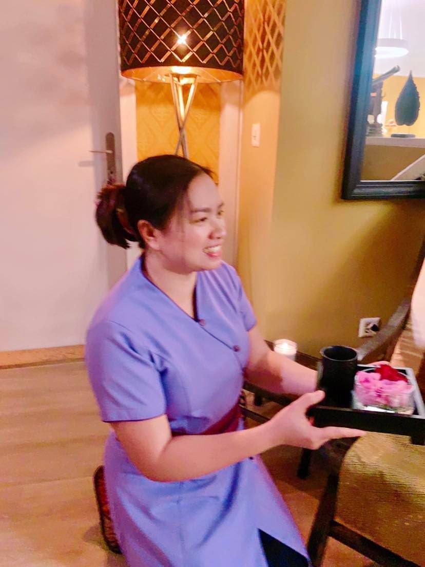 Frau in Lavendel-Peeling, kniend, mit einem Tablett mit Spa-Artikeln in der Hand, lächelnd in einem Spa-Ambiente.