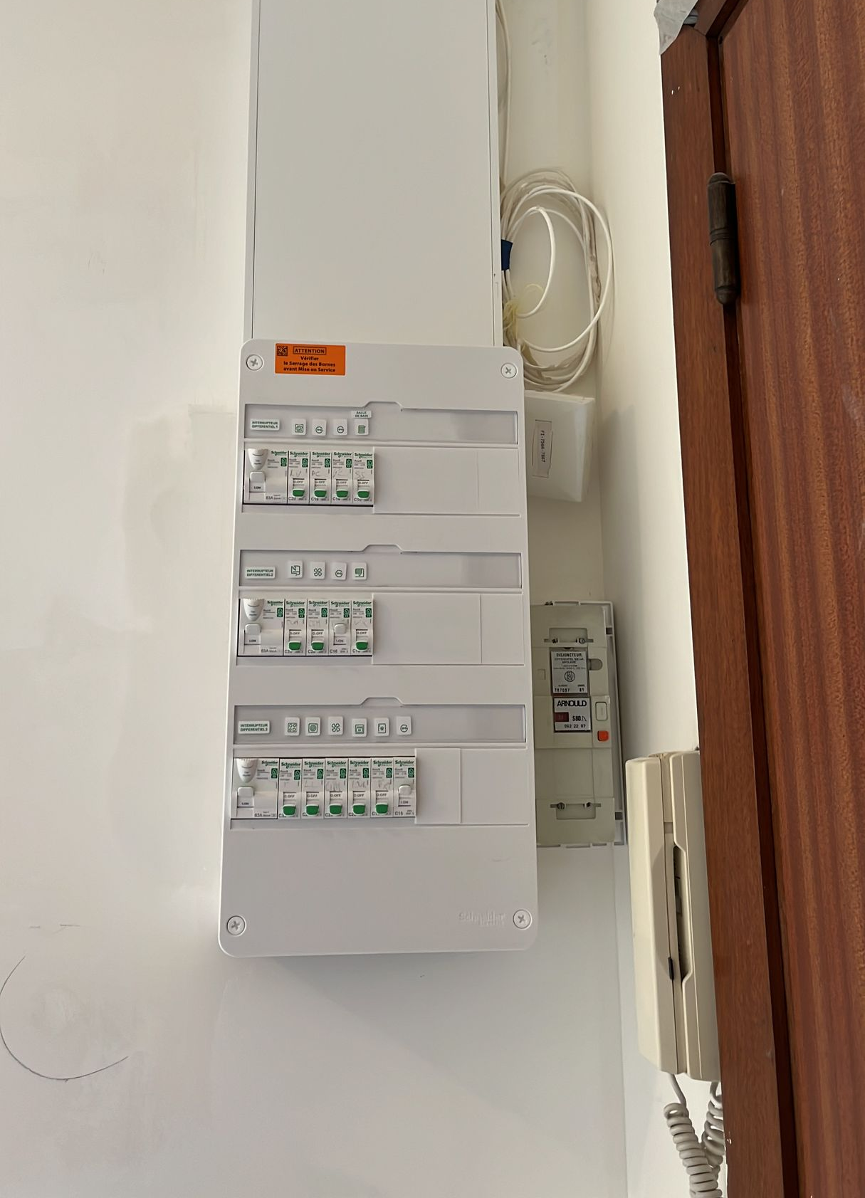 Tableau électrique blanc fixé au mur, avec divers interrupteurs et un compteur, à côté d'une porte marron.