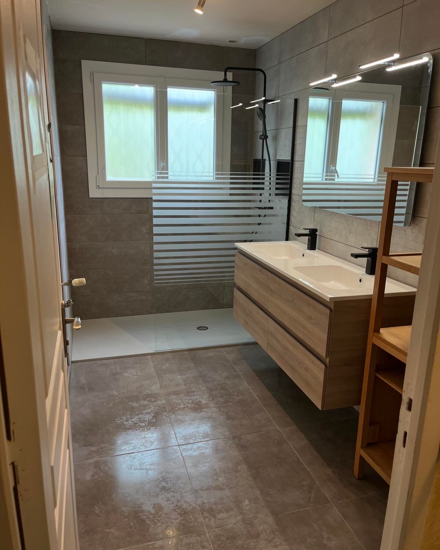 Salle de bains moderne avec double vasque, douche et fenêtre. Carrelage gris et touches de bois.