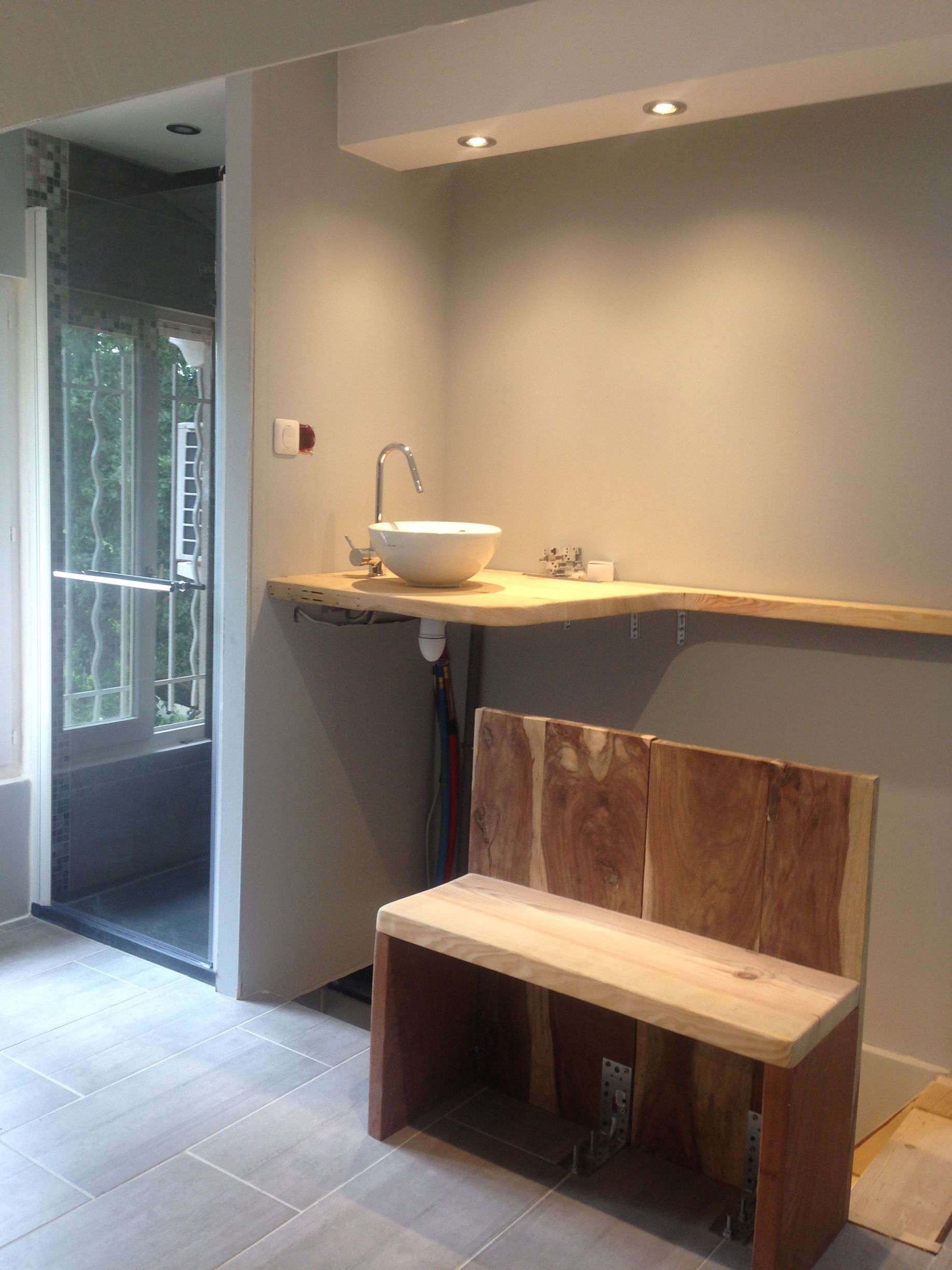 Salle de bains avec lavabo, banc et ascenseur. Comptoir en bois clair, lavabo blanc, murs gris et sol carrelé gris.