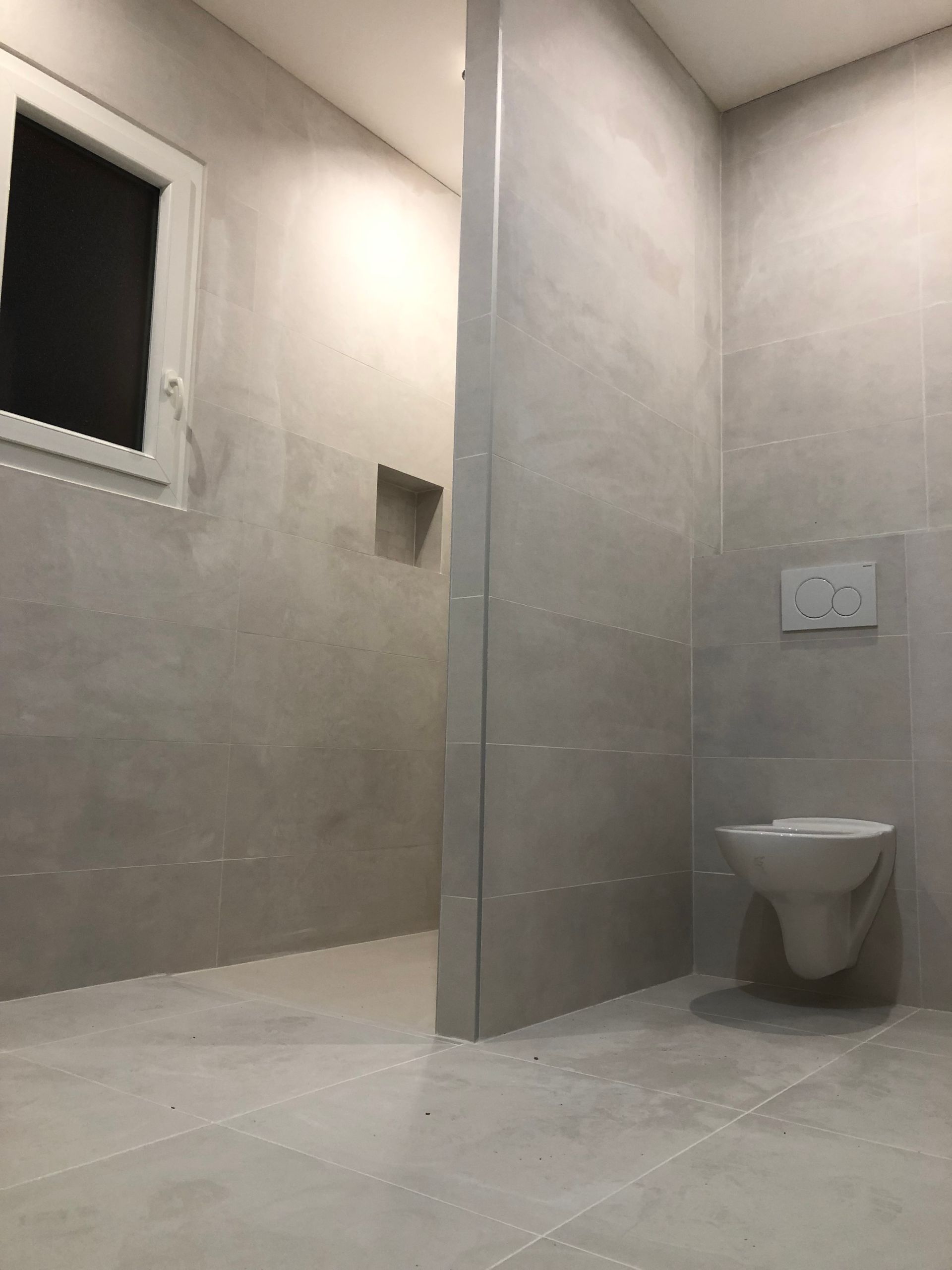 Salle de bains moderne avec carrelage gris, toilettes suspendues et fenêtre.