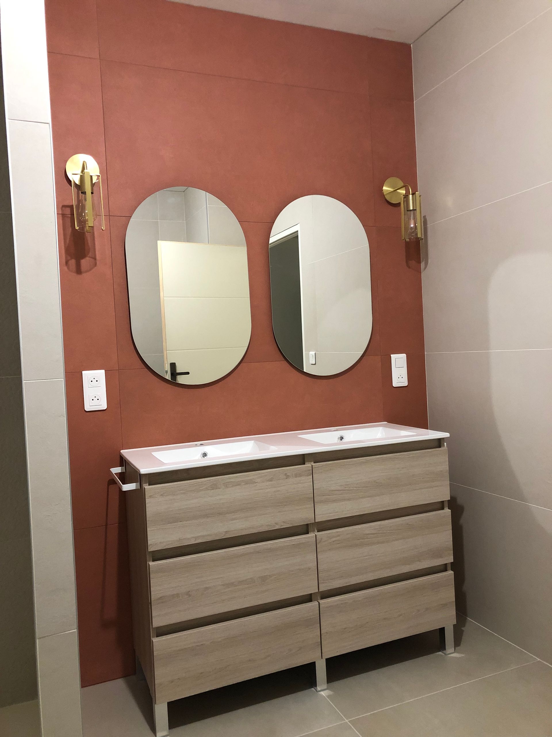 Salle de bains avec meuble-lavabo en bois, miroirs ovales et appliques murales sur un mur corail.