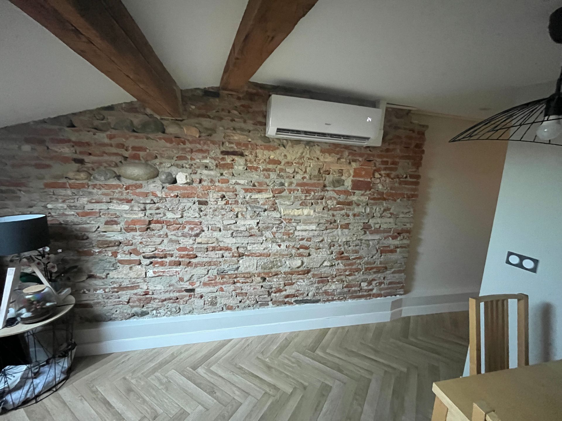 Intérieur d'une pièce avec mur en briques apparentes, climatiseur, poutres en bois et parquet à chevrons.