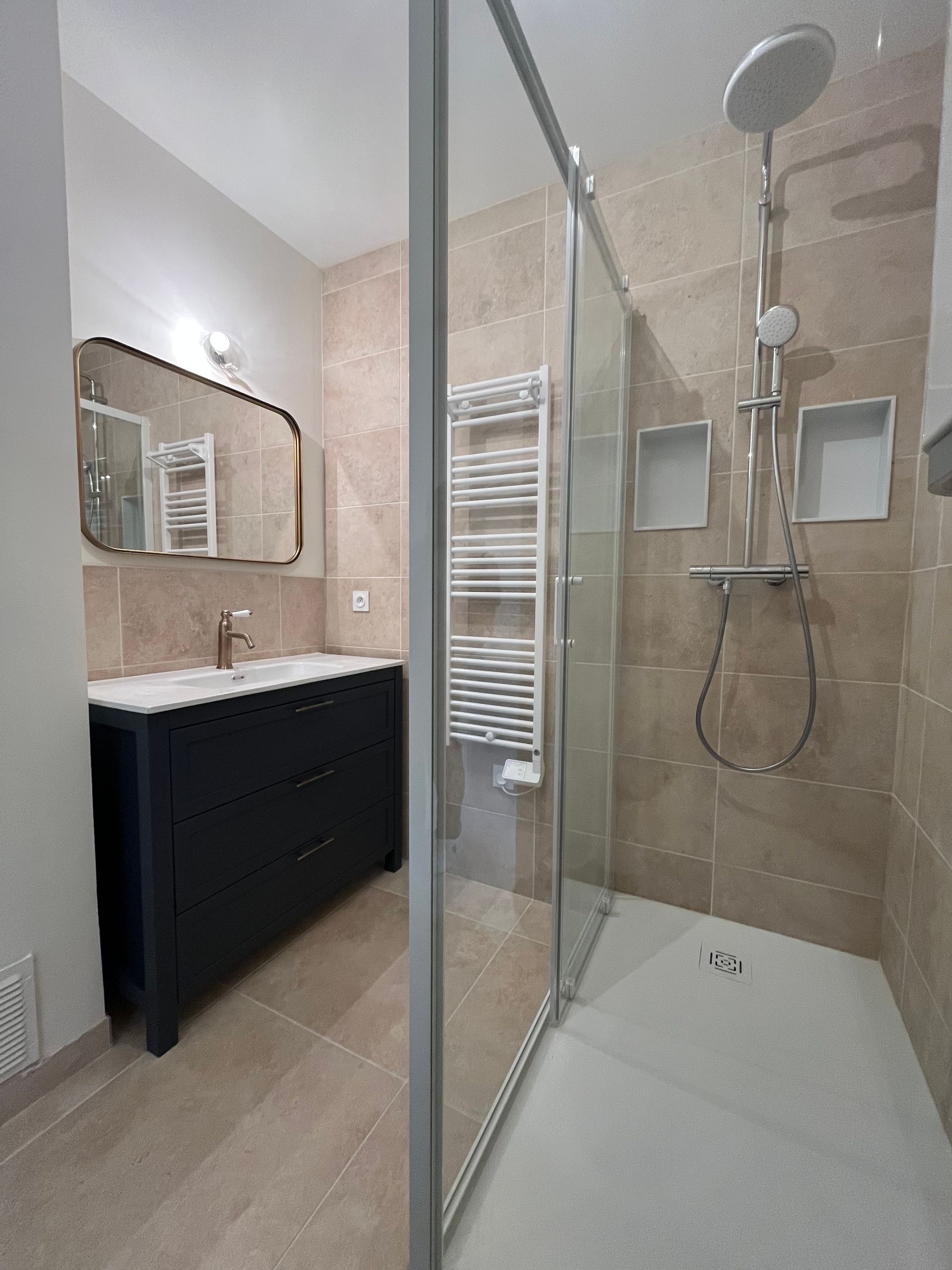 Salle de bains moderne avec douche, meuble-lavabo et miroir ; carrelage beige, meuble bleu marine et porte de douche en verre transparent.