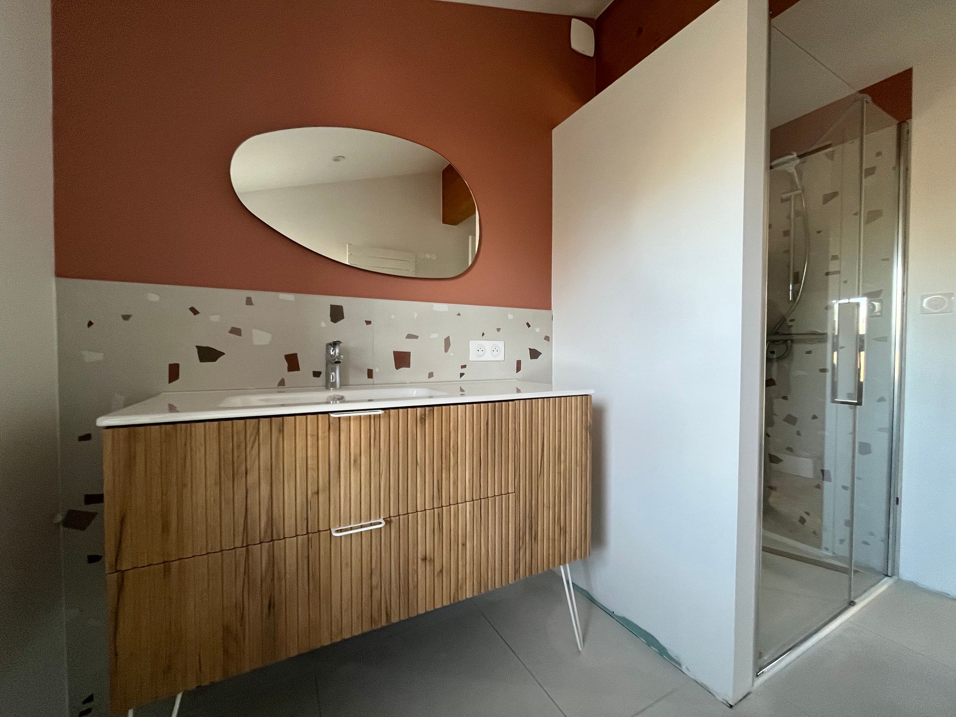 Salle de bains avec meuble-lavabo en bois, miroir ovale et douche en verre. Mur en terre cuite, cloison blanche.