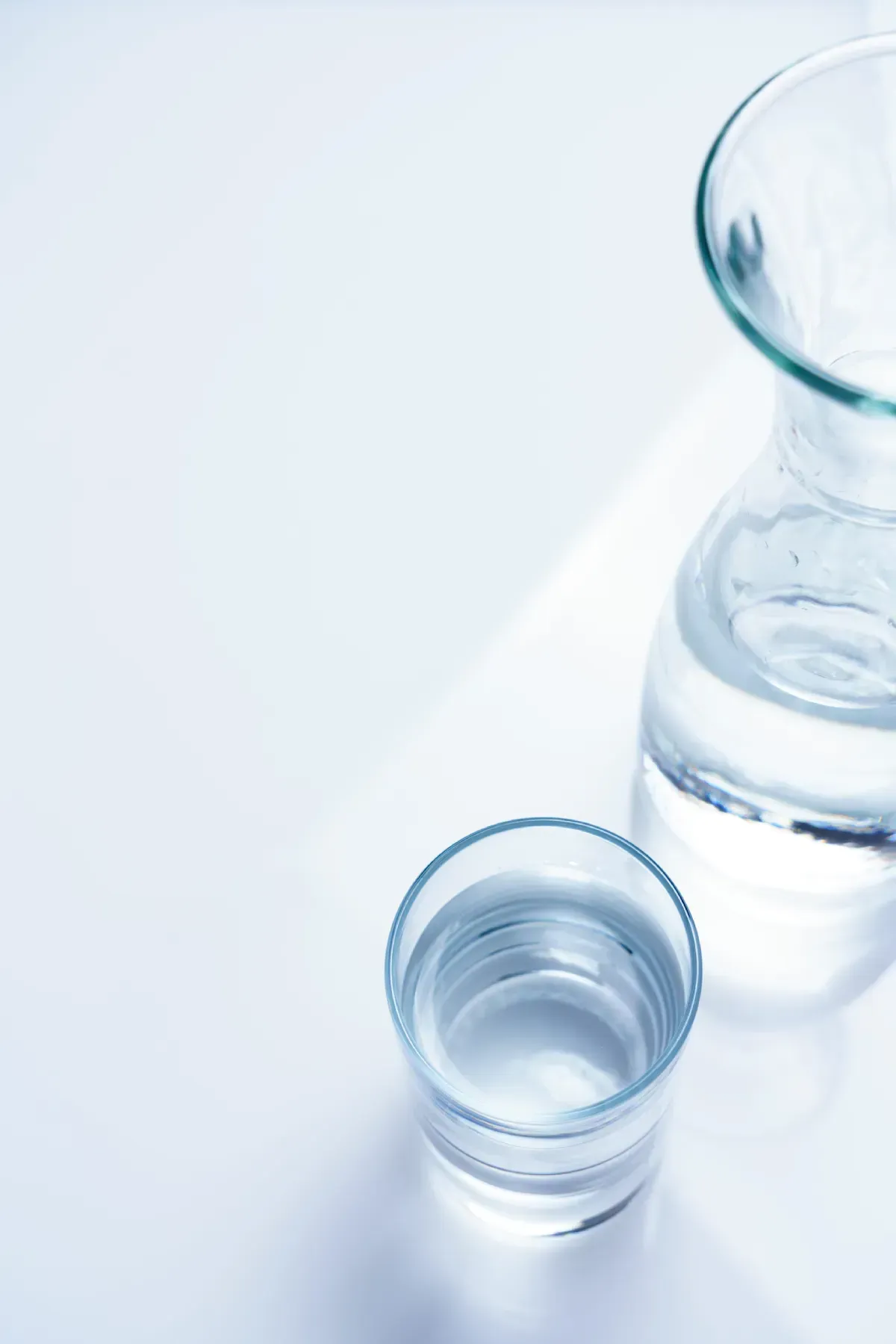 Un vaso de agua clara colocado junto a una jarra de agua a juego sobre una superficie blanca brillante y minimalista.