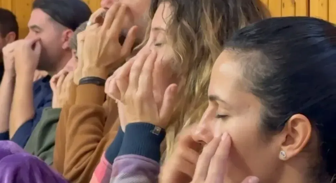 Un grupo de personas sentadas en fila con los ojos cerrados, presionando suavemente sus dedos contra sus rostros.
