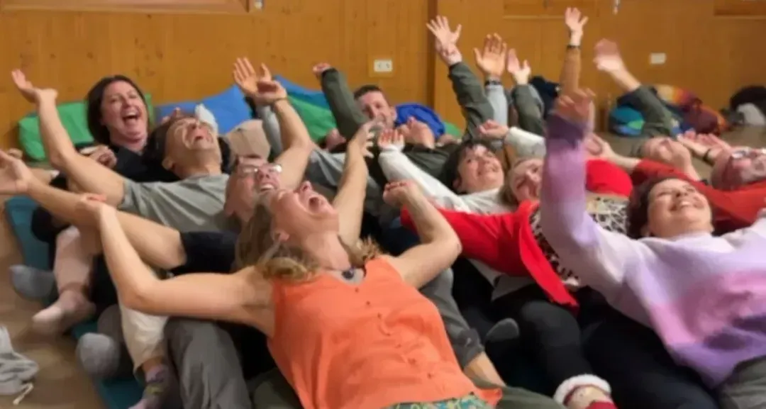 Un grupo de personas tumbadas en el suelo formando un círculo, con los brazos en alto, riendo y mirando hacia arriba