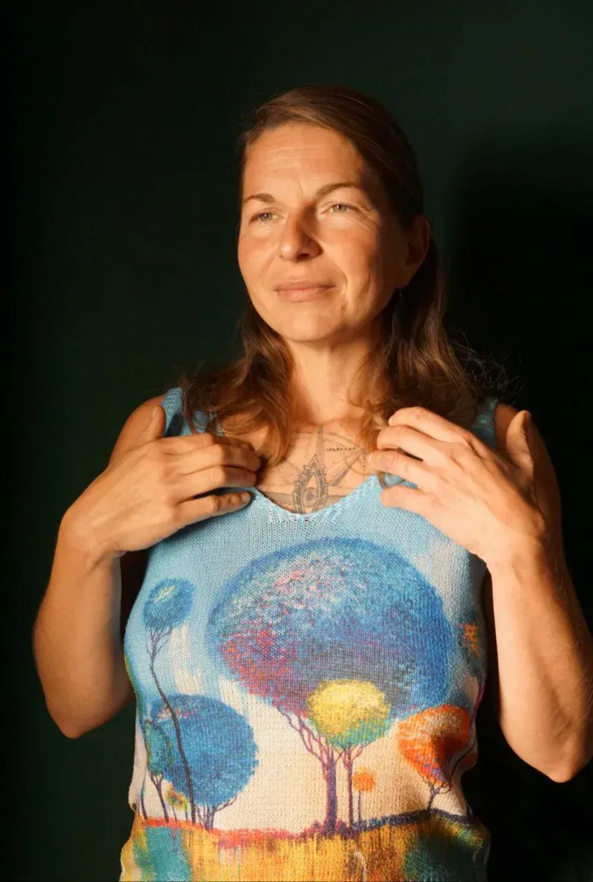 Una persona con cabello largo castaño posa frente a un fondo oscuro, vistiendo una blusa azul claro