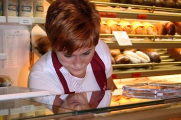 Frau mit roten Haaren in einer Bäckerei, die auf Backwaren hinter einer Glastheke hinunterblickt.