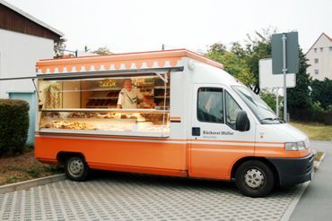 Orange-weißer Bäckerwagen, der Backwaren verkauft; eine Person steht hinter der Theke.