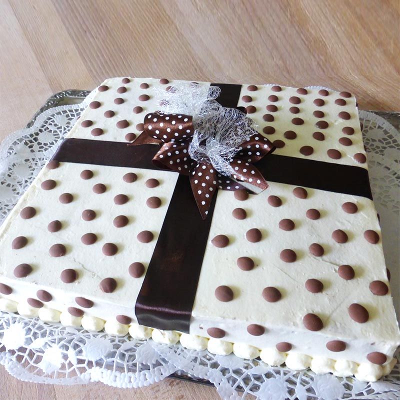 Quadratischer Kuchen, weißer Zuckerguss mit Schokoladenpunkten, braunes Band und Schleife, auf einem Deckchen.