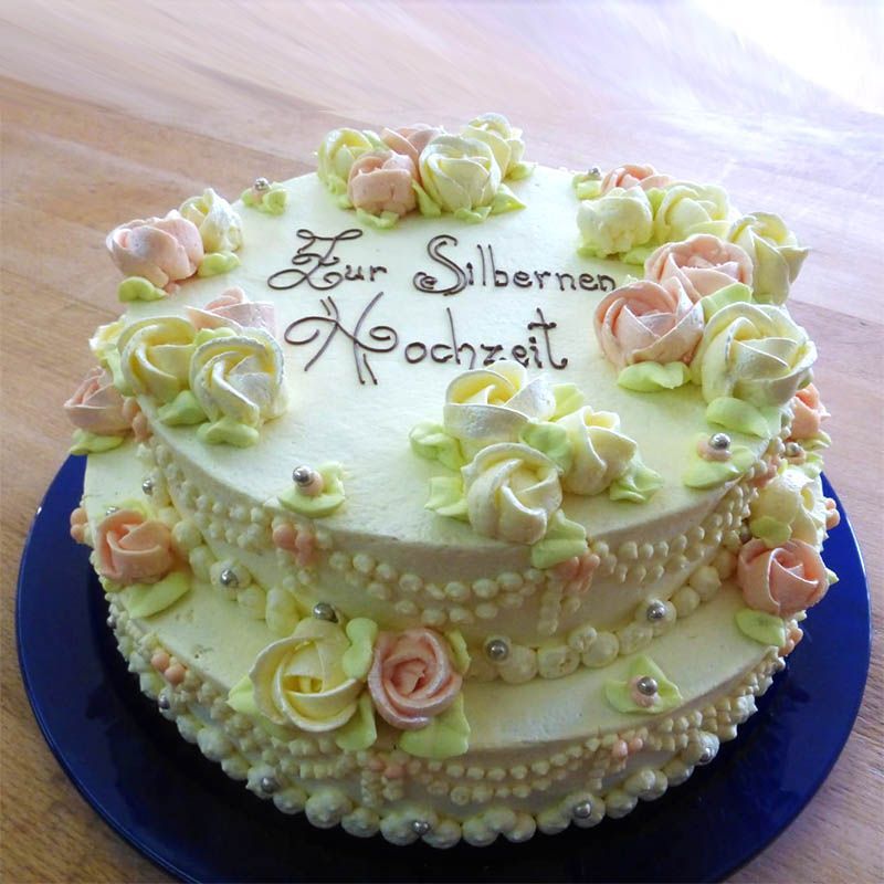 Zweistöckige Torte mit gelbem Zuckerguss, verziert mit aufgespritzten cremefarbenen Rosen in Gelb und Rosa und dem Text „Zur Silbernen Hochzeit“.