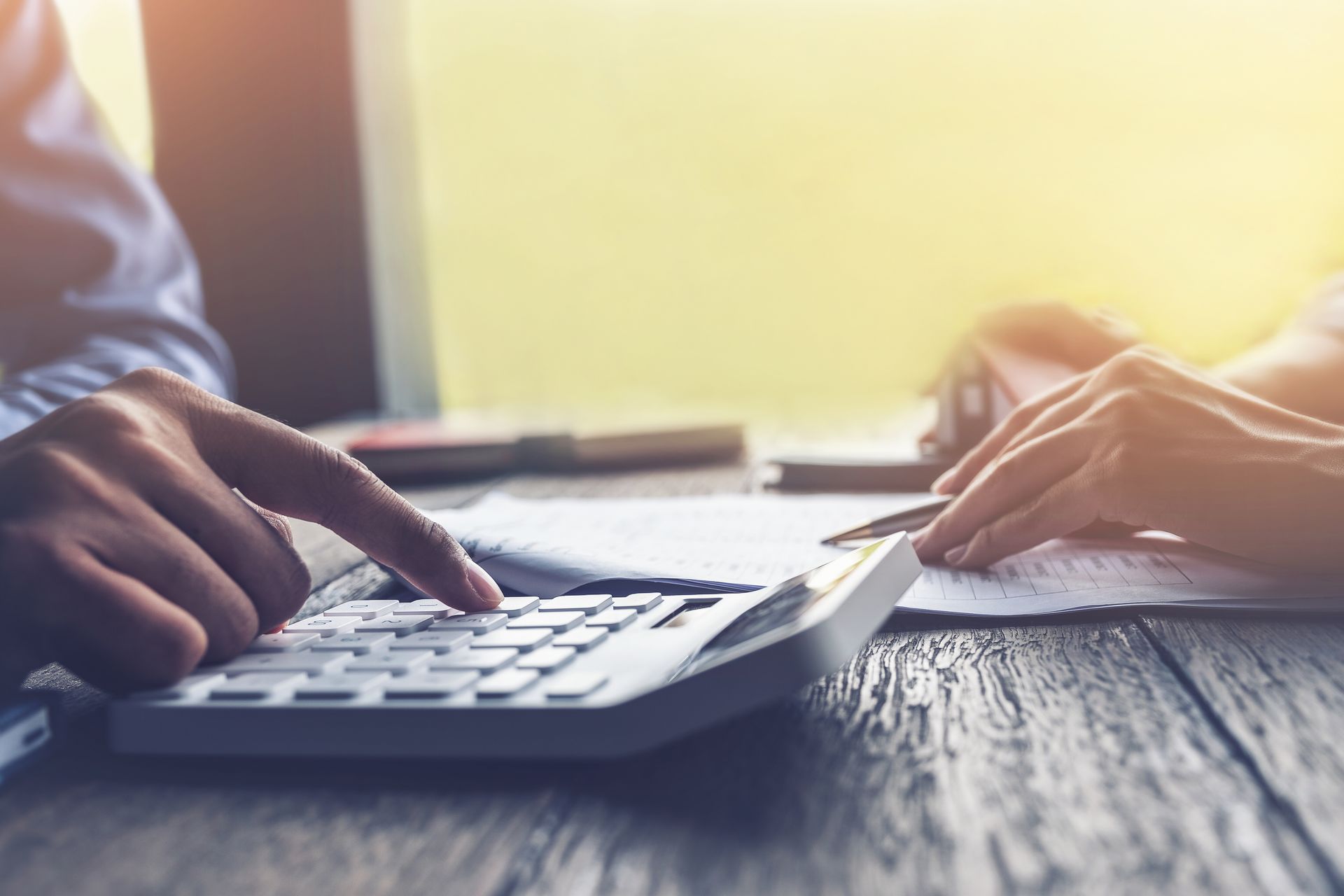 Des mains manipulant une calculatrice sur une table en bois, des papiers et un stylo à proximité ; lumière du soleil en arrière-plan.