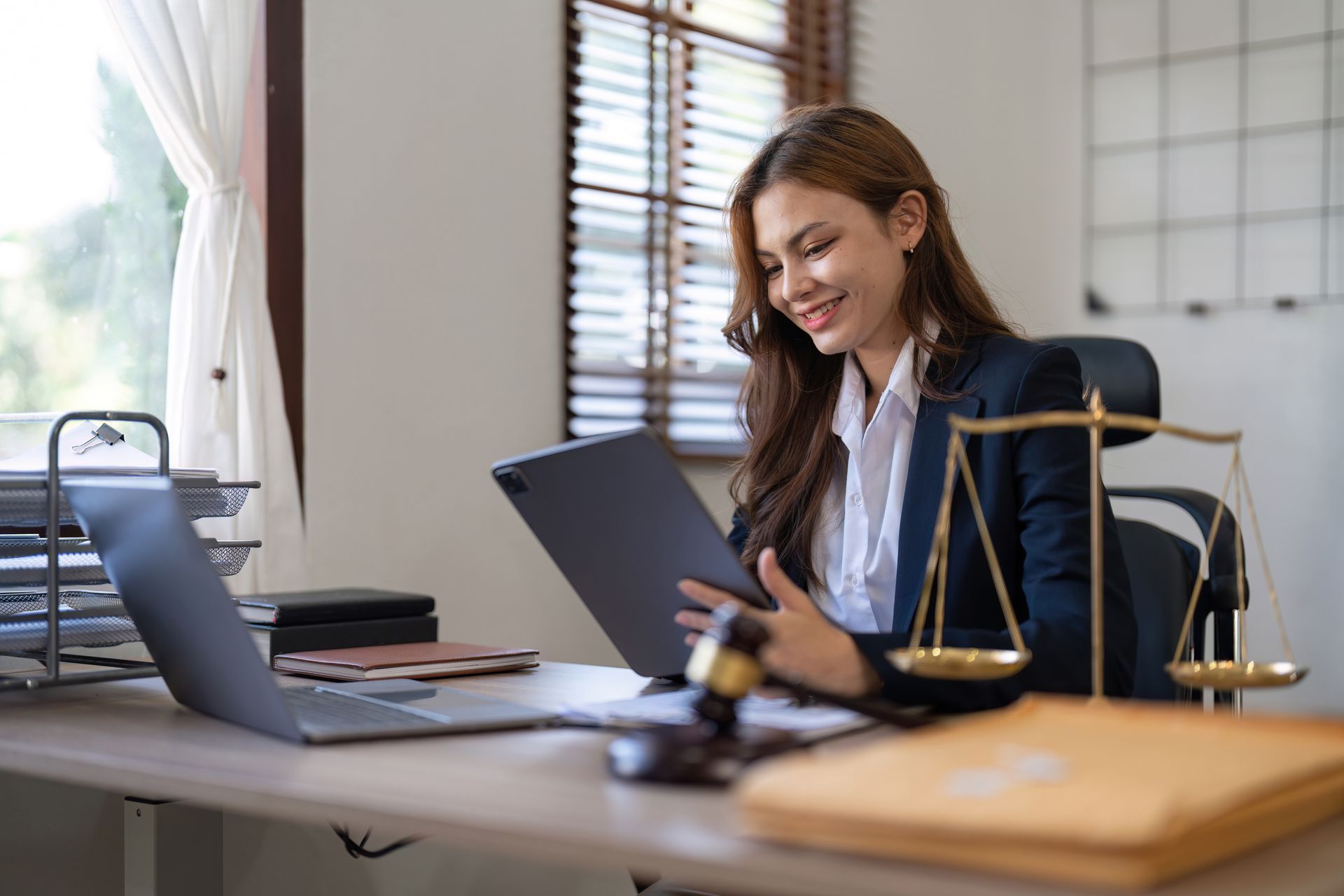 Une femme en blazer sourit en regardant une tablette sur un bureau où se trouvent un ordinateur portable, une balance de la justice et un marteau.