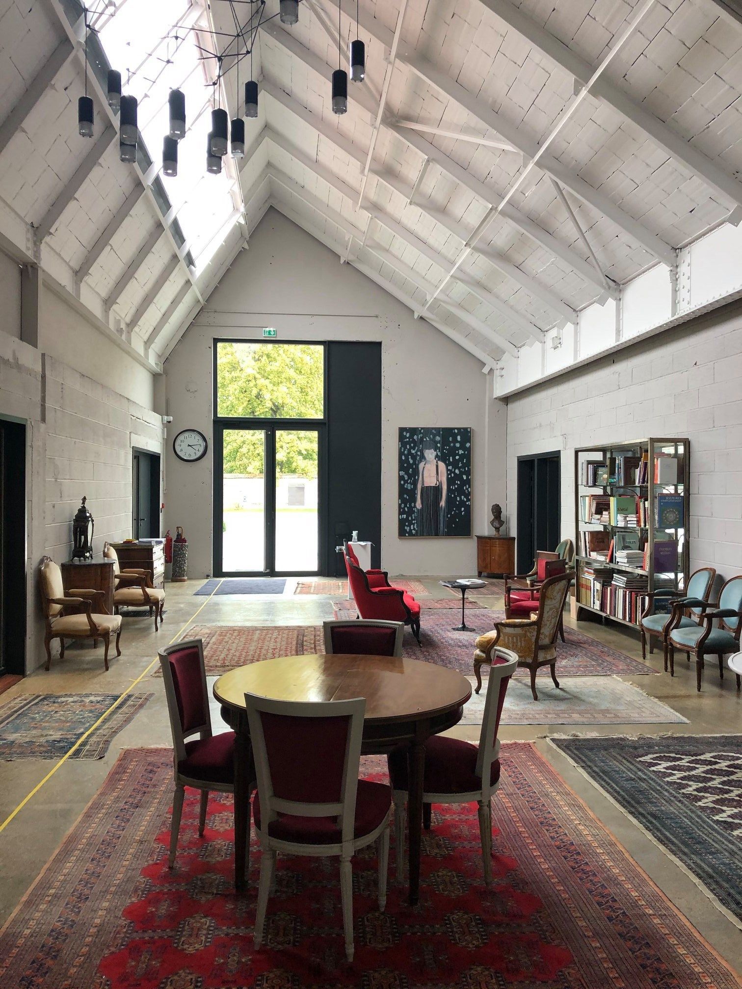 Salle d'exposition avec haut plafond blanc, tapis rouge, table ronde, chaises et bibliothèques près de grandes fenêtres