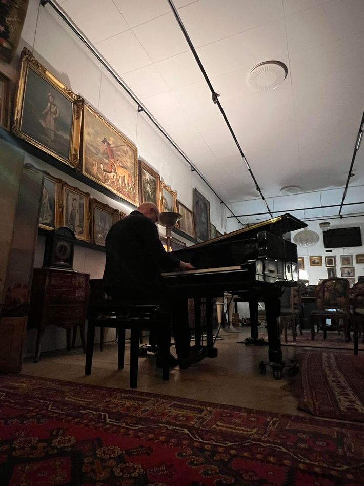 Un pianiste assis à un piano à queue dans une salle remplie d'œuvres d'art, avec des tapis et des tableaux ornementaux.