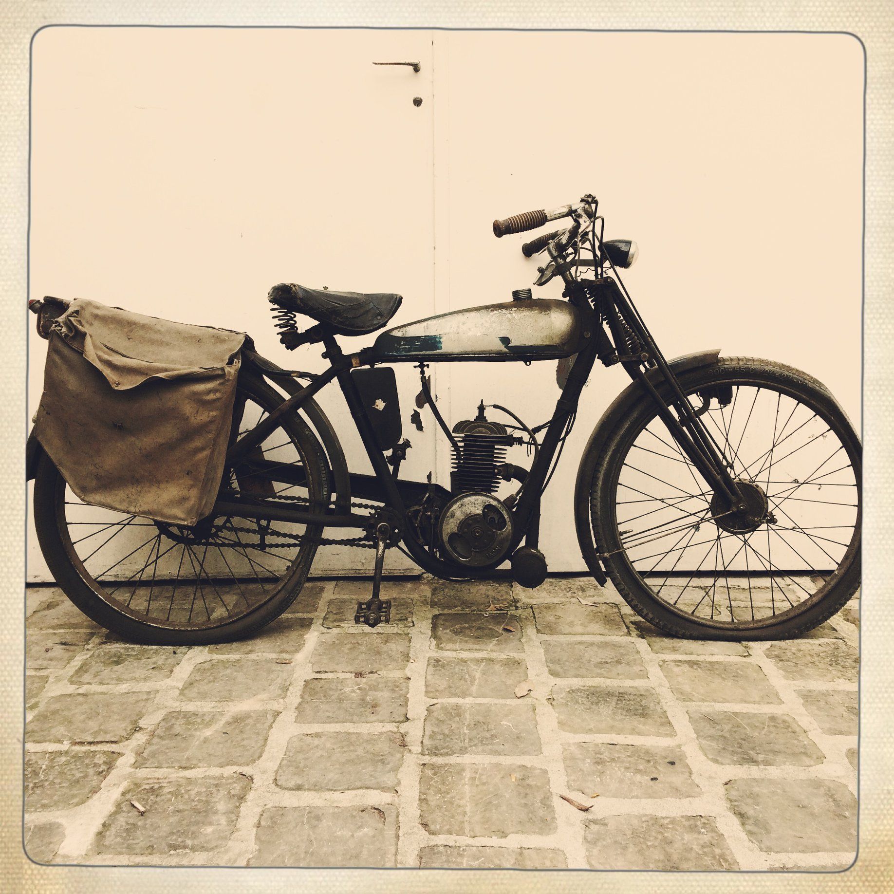 Moto vintage avec sacoche latérale beige, garée sur un sol en pierre contre un mur nu.