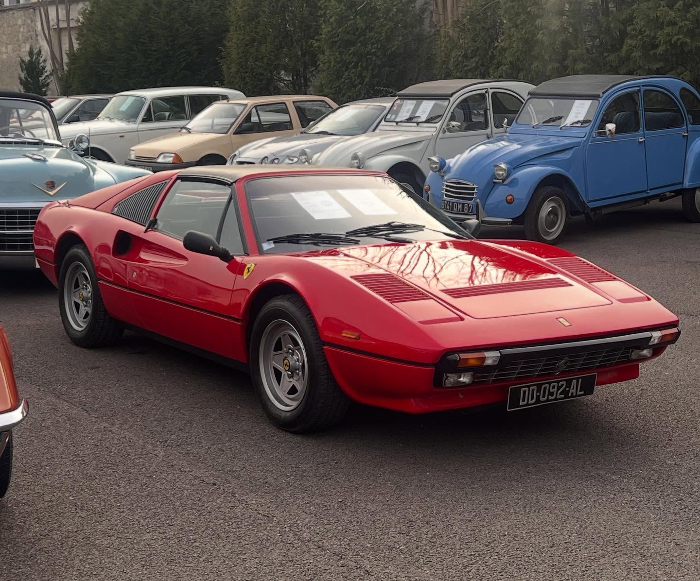 Voiture de sport rouge garée sur un parking, avec plusieurs voitures anciennes en arrière-plan.