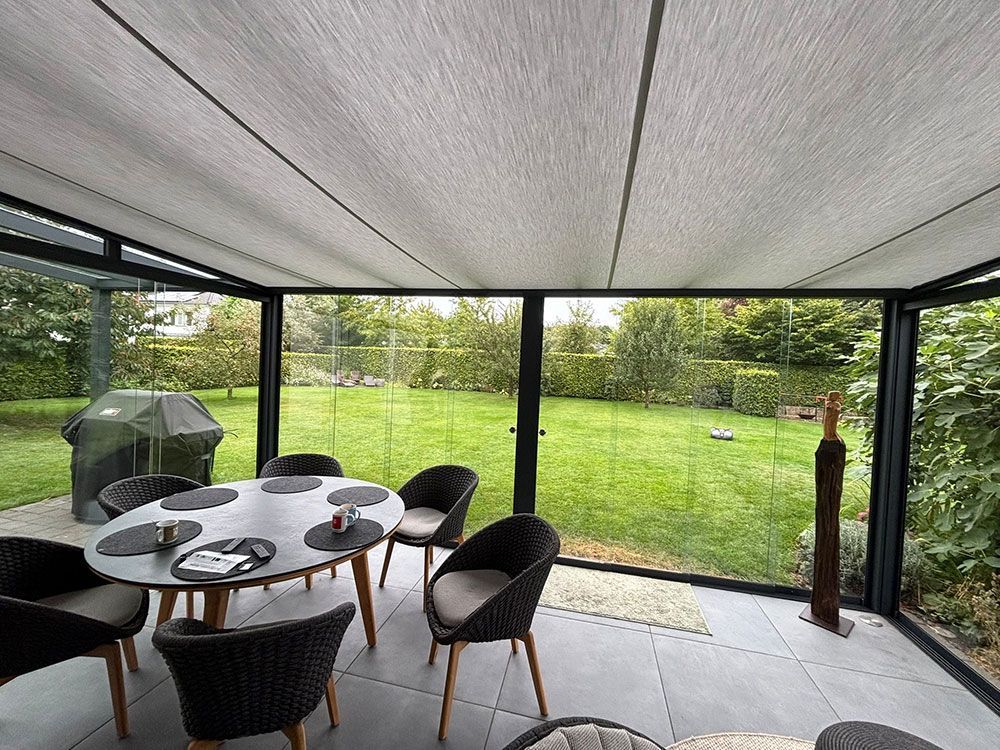 Wintergarten mit Glaswänden, grauem Dach, rundem Tisch, Stühlen und Blick auf einen grünen Garten.