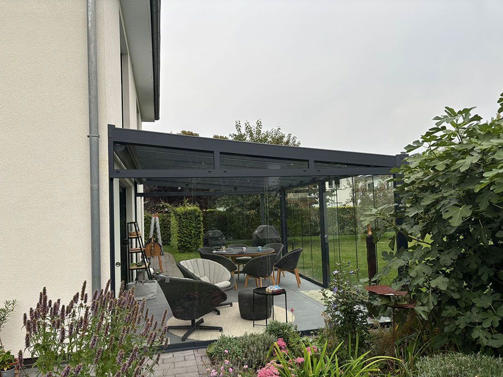 Terrasse mit dunklem Dach, Glaswänden und Sitzbereich neben einem Gebäude und Garten.