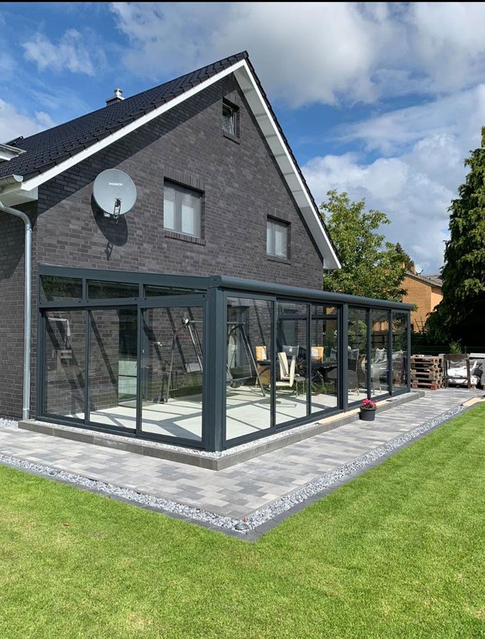 Wintergartenanbau mit grauen Zierelementen und Glaswänden an einem Backsteinhaus mit gepflasterter Terrasse.