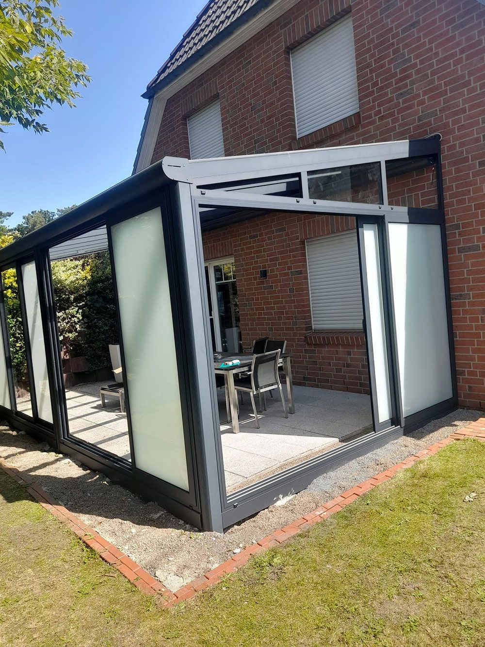Wintergarten mit Milchglasscheiben, der an ein Backsteinhaus angebaut ist. Im Inneren befinden sich ein Tisch und Stühle.