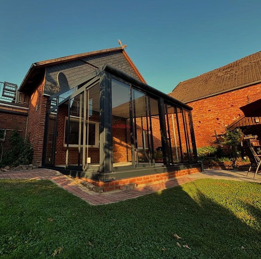 Wintergarten mit Glaswänden, an ein Backsteingebäude angebaut, mit einem Rasen davor.