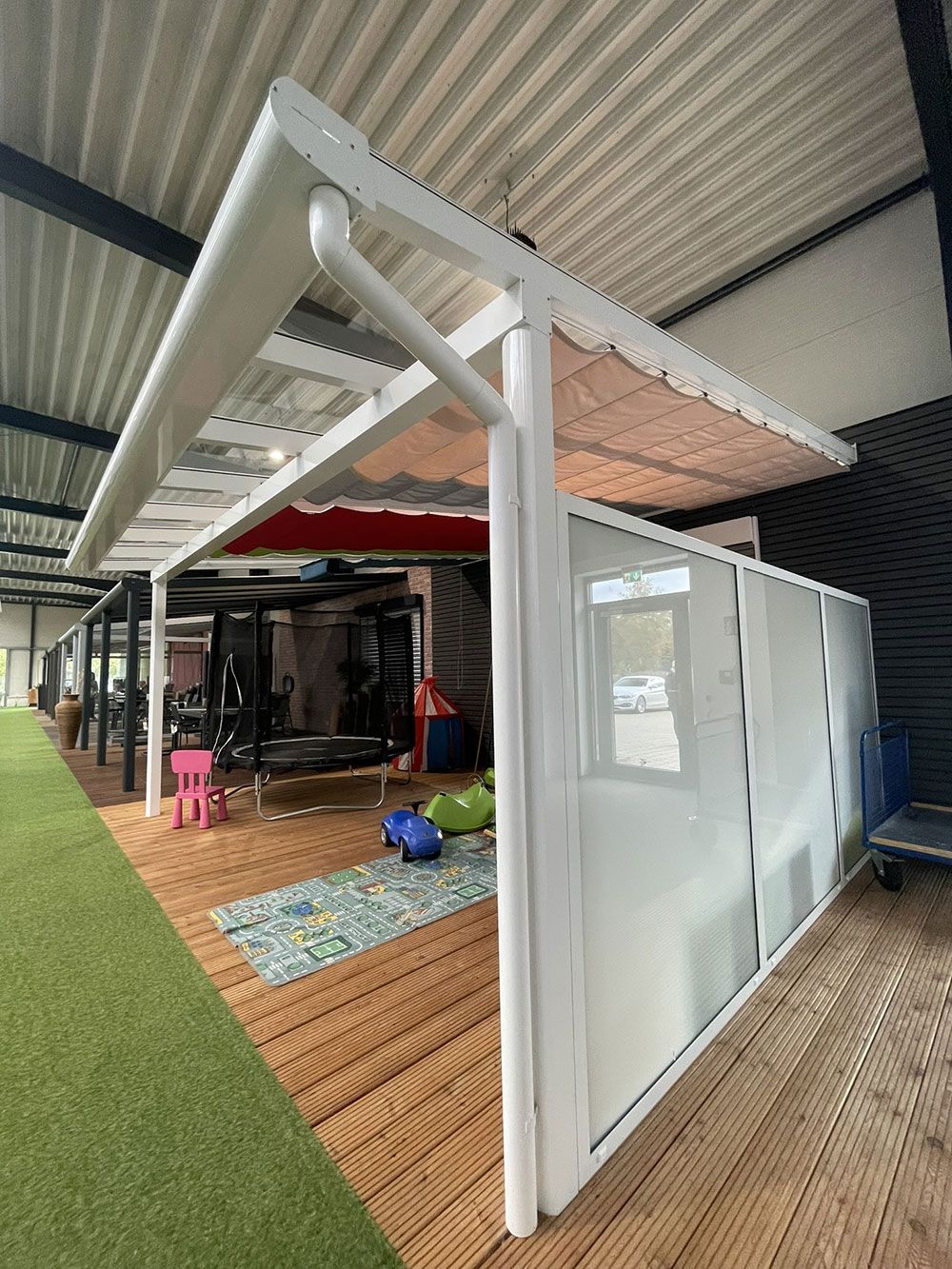 Überdachte Terrasse mit weißem Rahmen und beiger Markise, neben einem Gebäude.