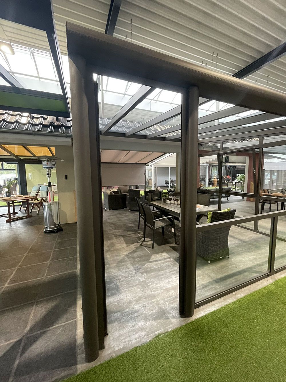 Essbereich im Freien mit Tischen, Stühlen und einer verglasten Terrasse unter einem Lamellendach.