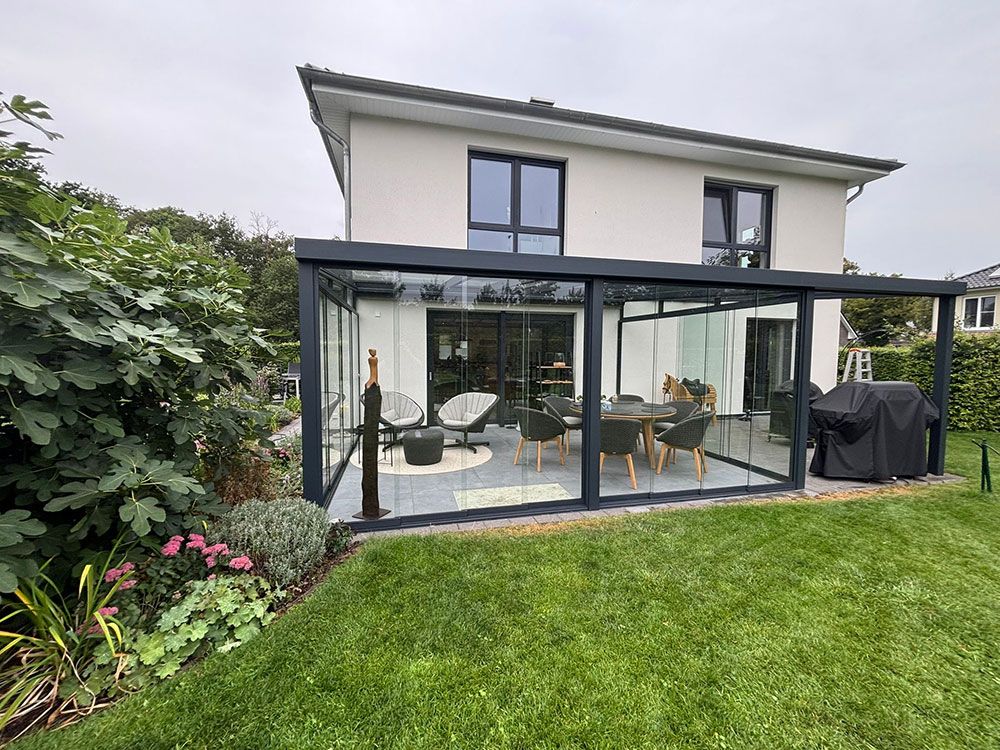 Verglaste Terrassenerweiterung an einem modernen Haus mit Gartenmöbeln und einem üppigen grünen Garten.