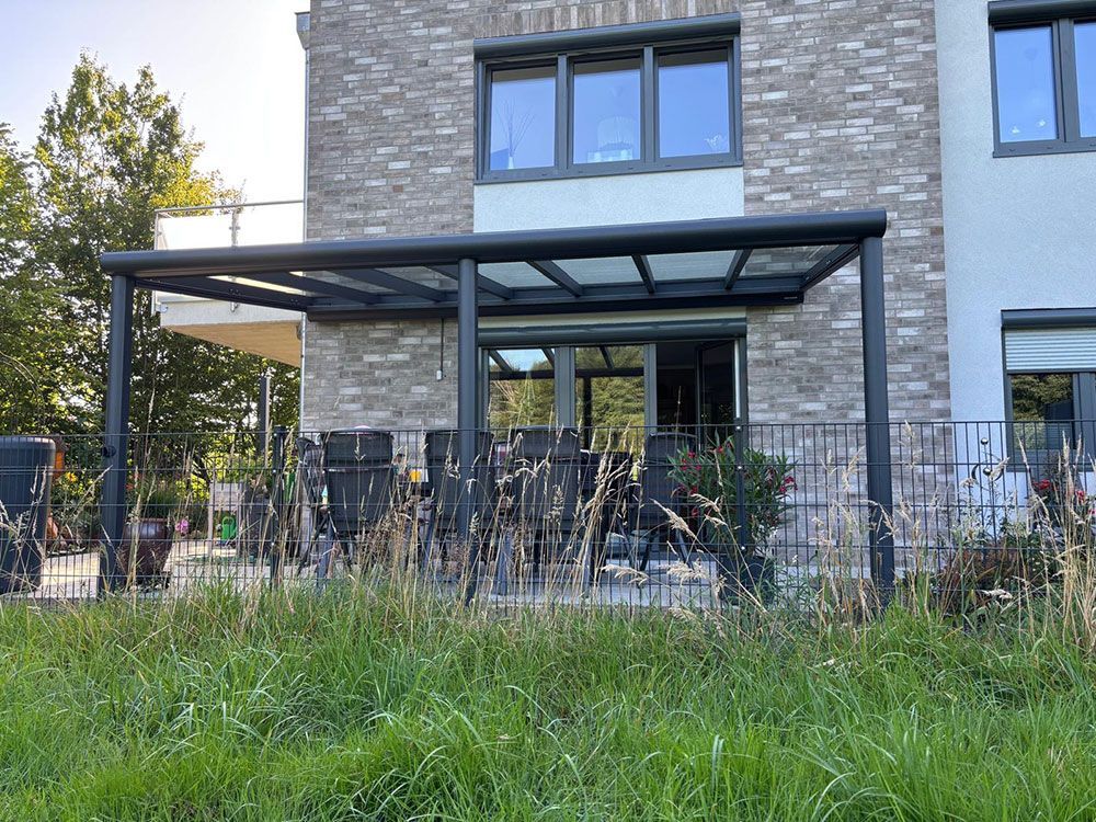 Schwarze Pergola über einer Terrasse mit Essmöbeln. Hohes Gras im Vordergrund, Backsteinhaus im Hintergrund.