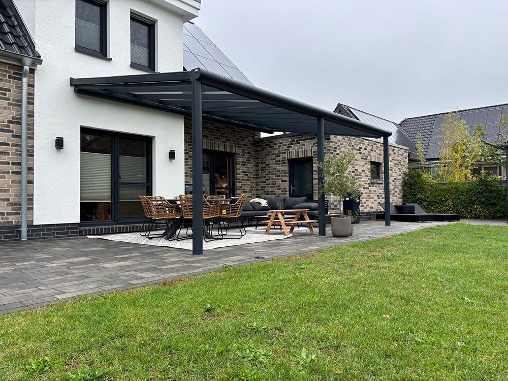 Moderne Terrasse mit dunkelgrauer Pergola, Backstein- und weißem Haus, Rasen und Gartenmöbeln.