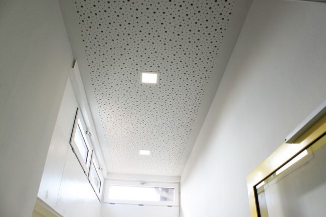 Detailansicht einer weißen Wand mit Fensterrahmen und perforierter Akustikdecke.