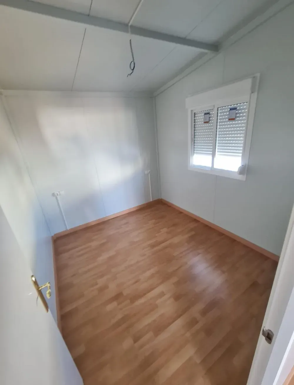 Habitación vacía con piso de madera clara, una ventana con persianas y paredes de color verde pálido.