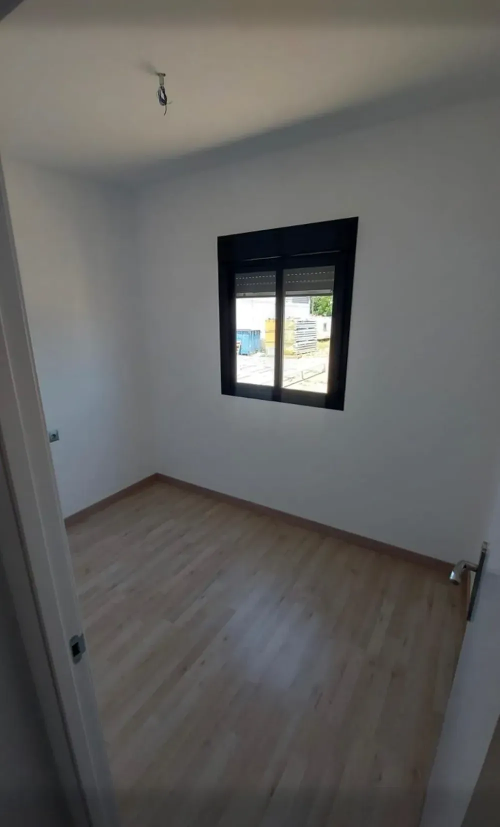 Habitación vacía con paredes blancas, piso de madera y pequeña ventana con marco negro.